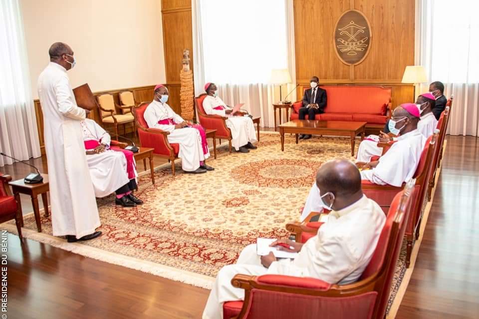 Huit hommes vêtus d’habits religieux et formels sont assis en cercle dans un salon orné d’un tapis et de boiseries avec un blason au mur