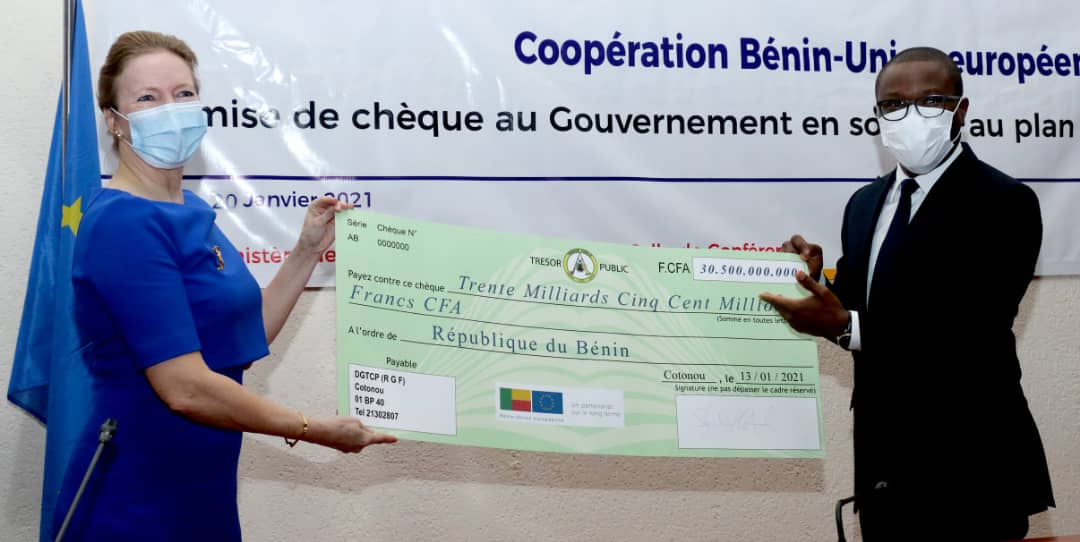 Deux personnes masquées tiennent un grand chèque représentant un don financier officiel devant une bannière de coopération internationale
