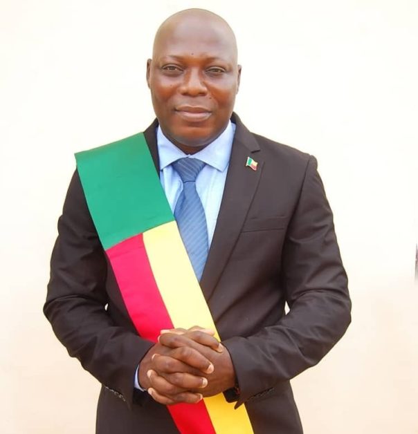 Homme en costume sombre avec une écharpe tricolore vert, jaune et rouge posant devant un fond blanc
