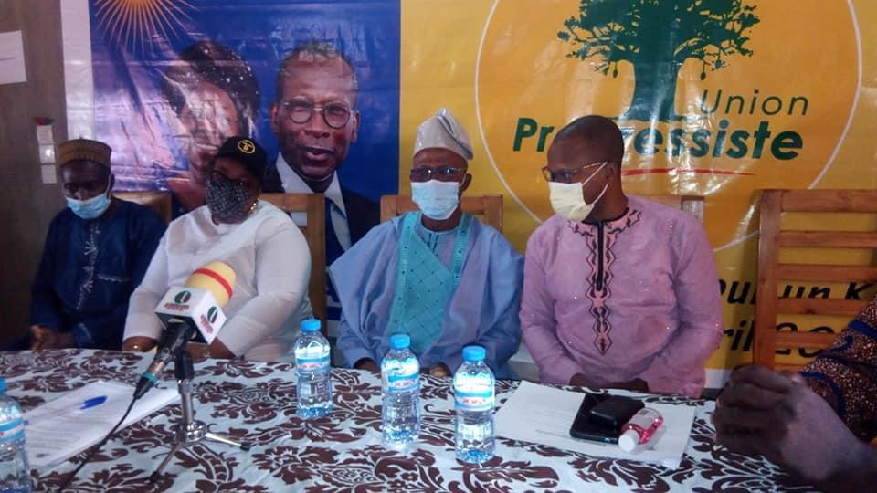 Quatre hommes masqués assis à une table devant des banderoles et plusieurs bouteilles d'eau sur la table