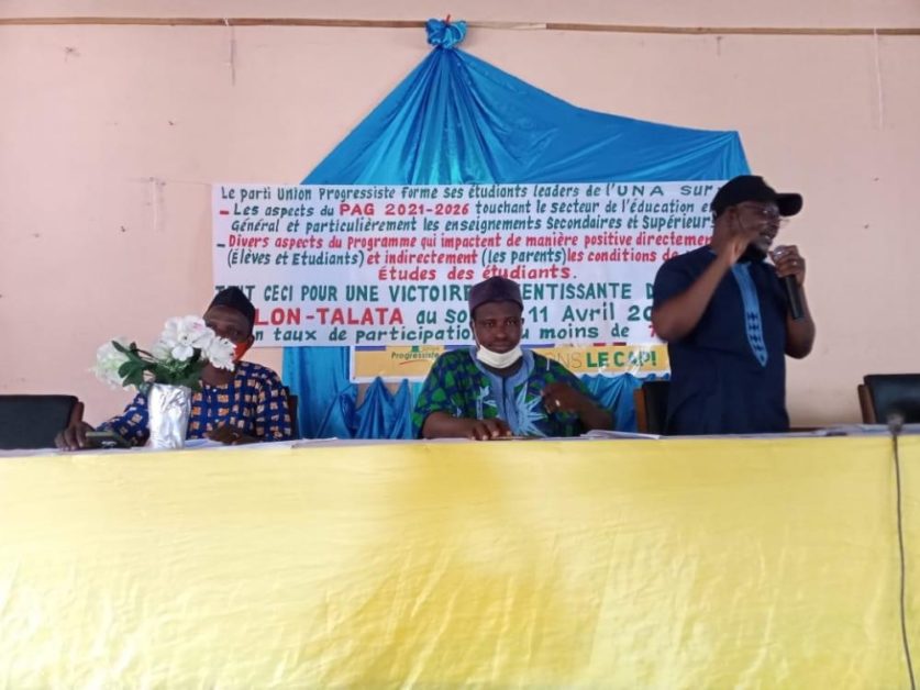 Trois hommes assis à une table avec microphone et pancarte bleue et blanche en arrière-plan