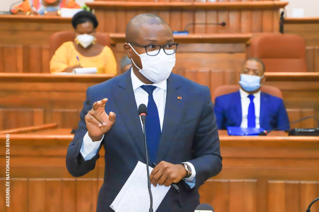 Homme en costume cravate et masque parlant debout dans une salle d’assemblée avec des personnes assises en arrière-plan