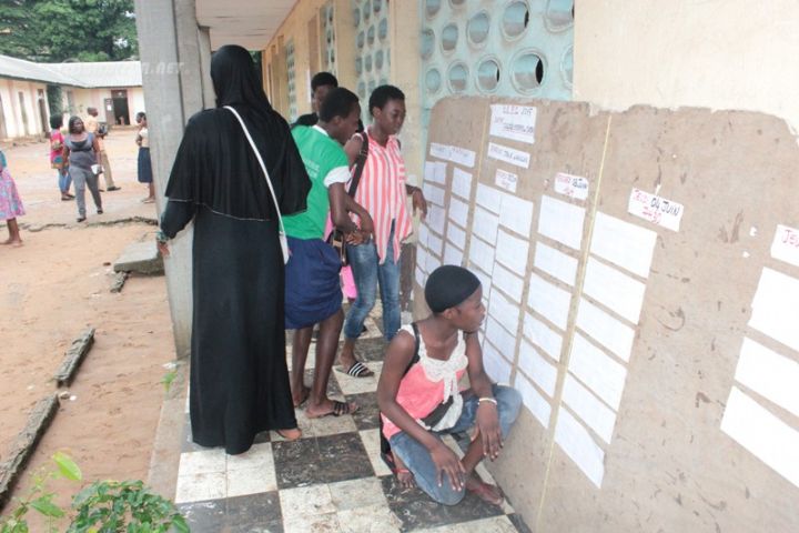Un groupe de jeunes consulte des listes affichées sur un mur à l'extérieur d'un bâtiment scolaire.