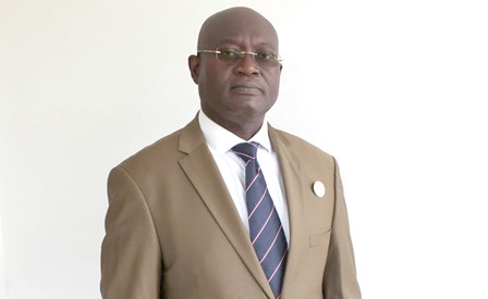 Homme en costume marron clair, cravate rayée, portant des lunettes, posé devant un mur blanc
