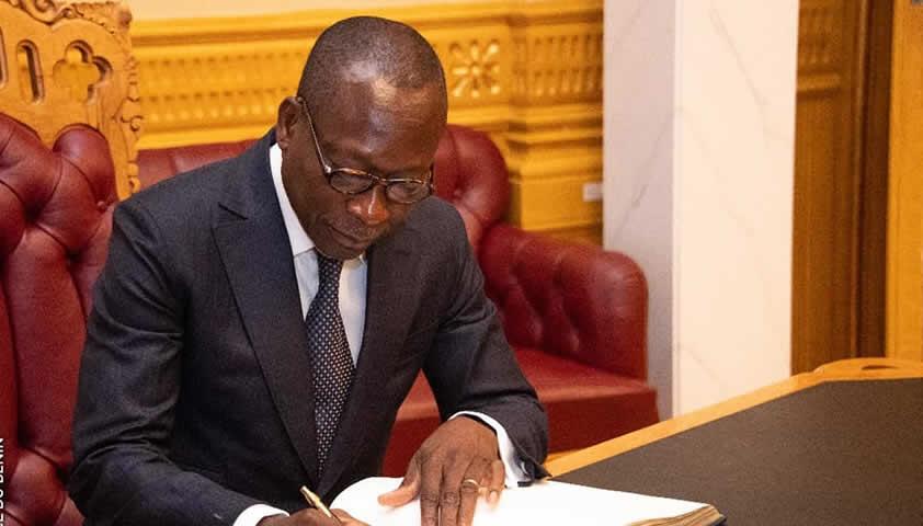Un homme en costume sombre et lunettes signe un document derrière un bureau dans un intérieur luxueux.