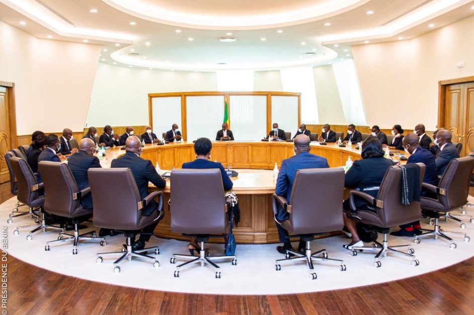 Personnes assises autour d'une grande table ovale en réunion dans une salle lumineuse avec un drapeau au centre