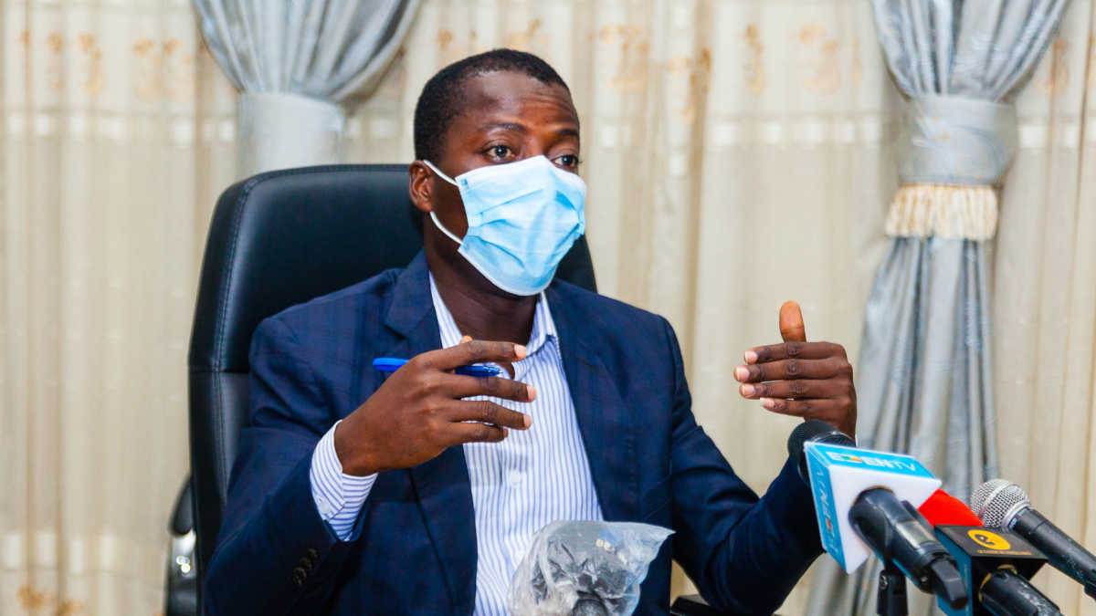 Un homme portant un masque bleu parle et gesticule assis derrière un bureau avec plusieurs micros devant lui