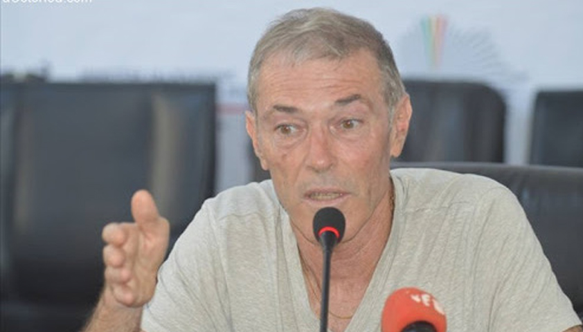 Homme en chemise claire parlant devant un micro lors d’une conférence avec un fond flou et un autre micro rouge devant lui
