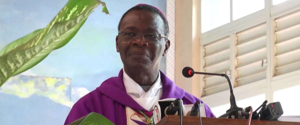 Un homme en tenue religieuse violette parle derrière un pupitre avec micro dans une pièce éclairée par une fenêtre blanche