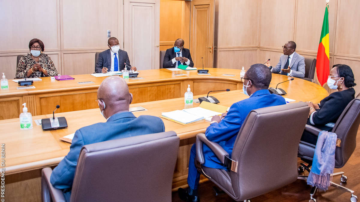 Sept personnes masquées assises autour d'une table de réunion avec un drapeau en arrière-plan