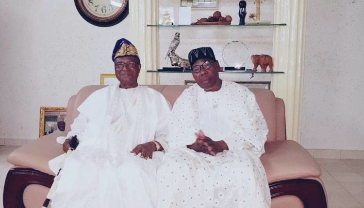 Deux hommes âgés en tenue traditionnelle africaine blanche assis sur un canapé devant une vitrine avec objets décoratifs