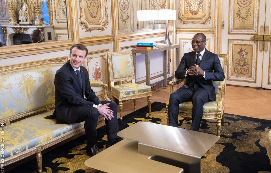 Deux hommes en costume discutent en souriant assis dans un salon richement décoré avec meubles dorés et tapis noir et or