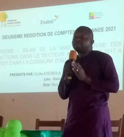 Un homme en tenue sombre parle devant un écran de projection affichant un texte en français sur une reddition de comptes