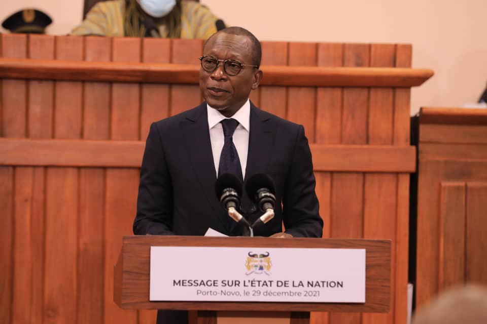 Un homme en costume foncé parle à un pupitre avec un panneau sur l’état de la nation devant un banc en bois dans une salle officielle
