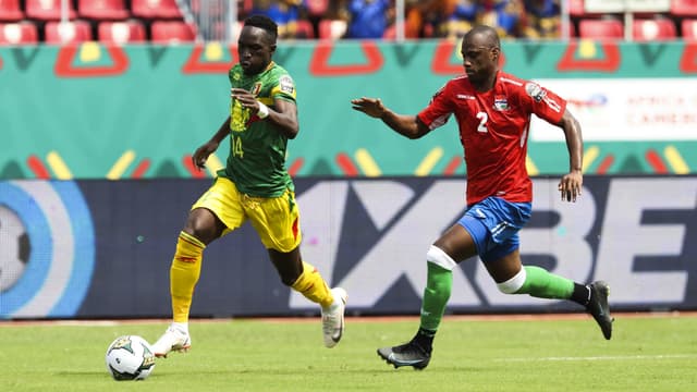 Un joueur en maillot vert et jaune contrôle le ballon devant un adversaire en maillot rouge et bleu sur un terrain de football en plein match