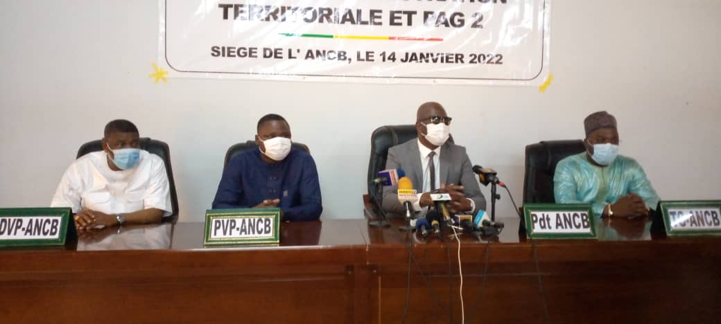 Quatre hommes portant des masques discutent à une table de conférence avec des pancartes indiquant leurs fonctions devant eux