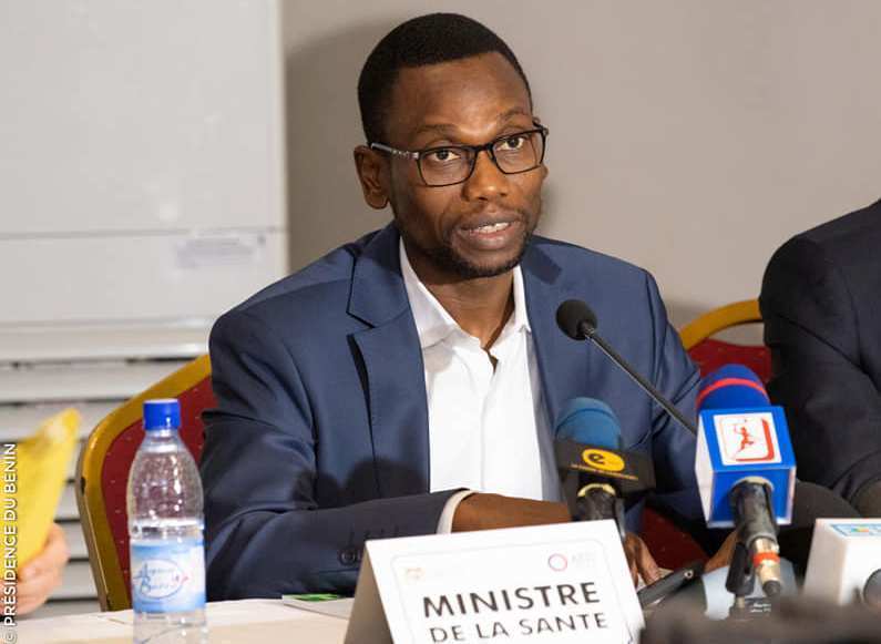 Un homme en costume bleu parle au micro lors d'une conférence avec des micros et une pancarte "Ministre de la Santé" devant lui