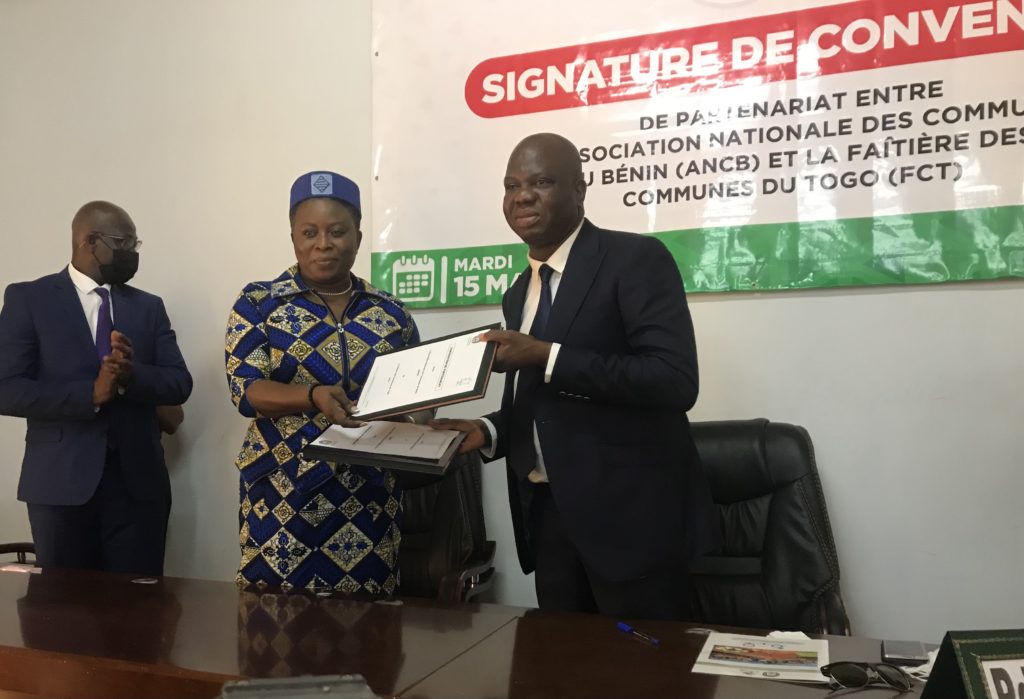 Deux personnes échangent des documents devant un panneau indiquant la signature d’une convention, un homme applaudit à gauche