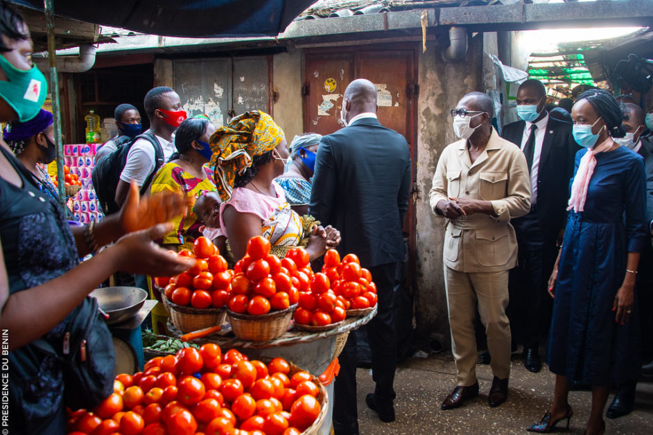 Un groupe de personnes masquées visite un marché avec des paniers de tomates et des produits alimentaires visibles
