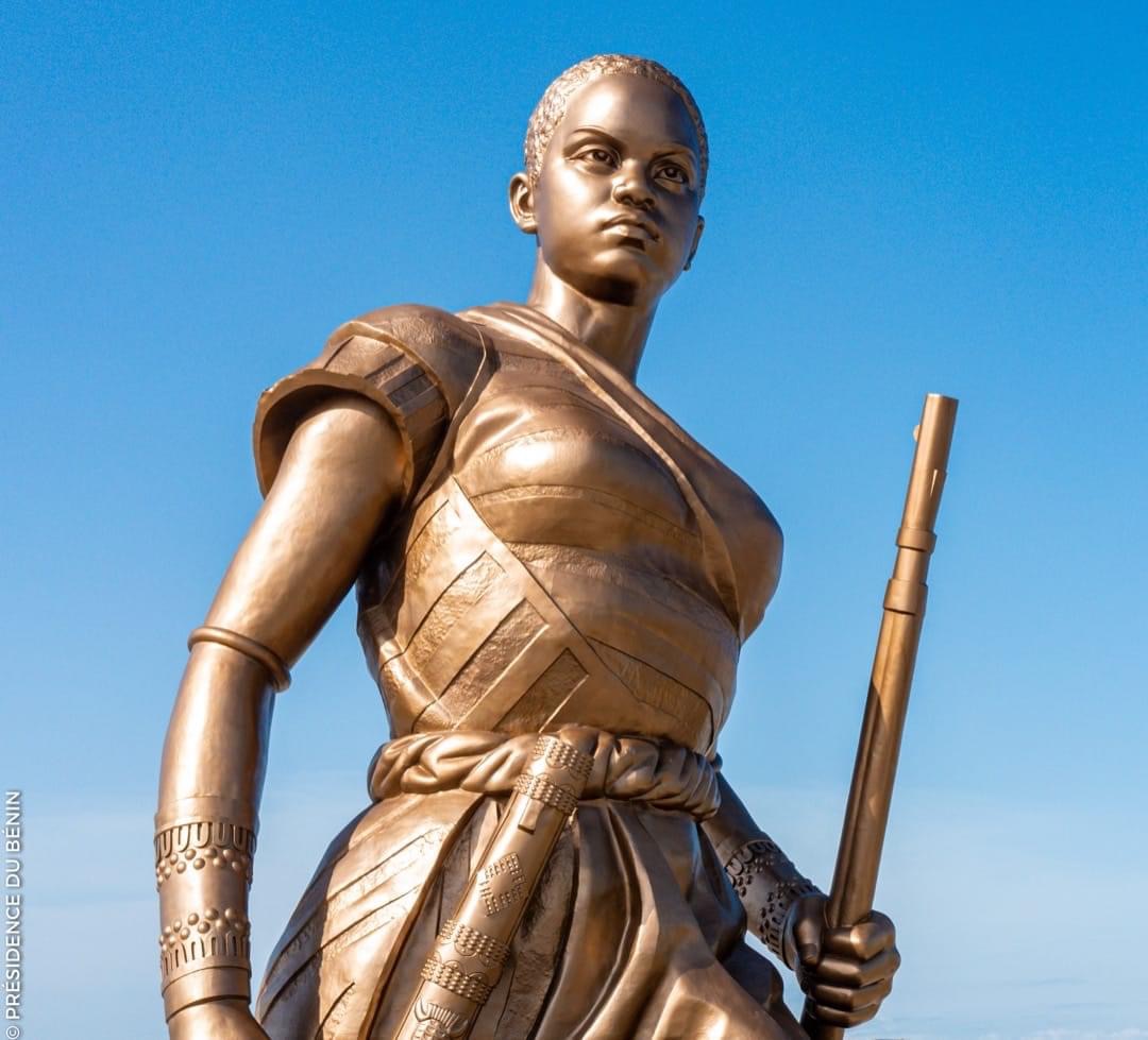 Statue en bronze d’une femme guerrière tenant un bâton, vêtue d’une tenue traditionnelle, sous un ciel bleu clair