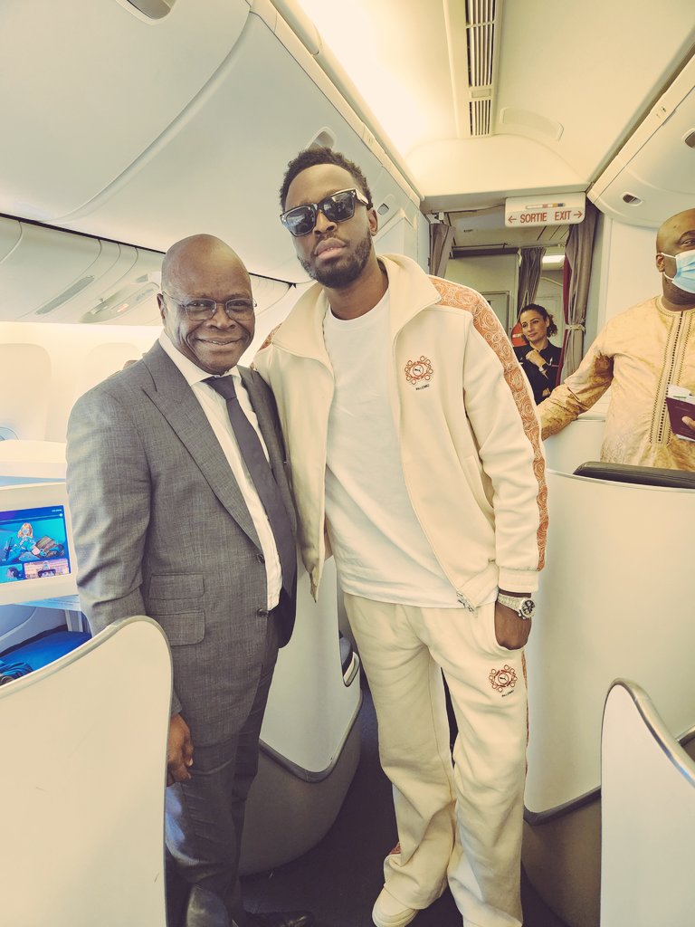 Deux hommes debout dans l'allée d'un avion, l'un en costume, l'autre en tenue décontractée avec lunettes de soleil, souriant