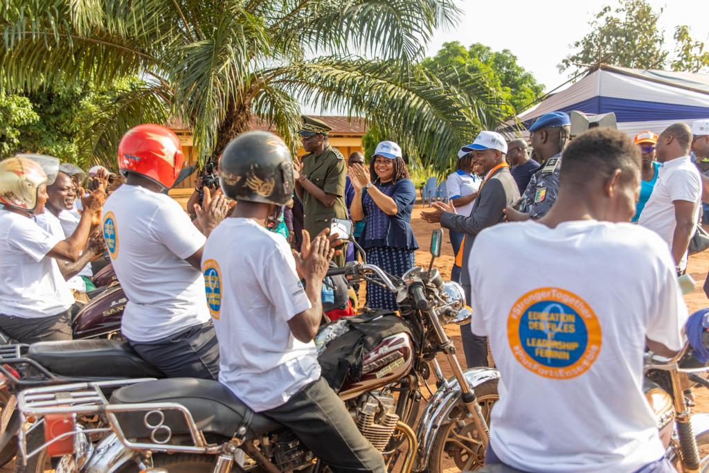 Groupe de motards en t-shirts blancs salués par un public sous des palmiers lors d'un rassemblement en plein air