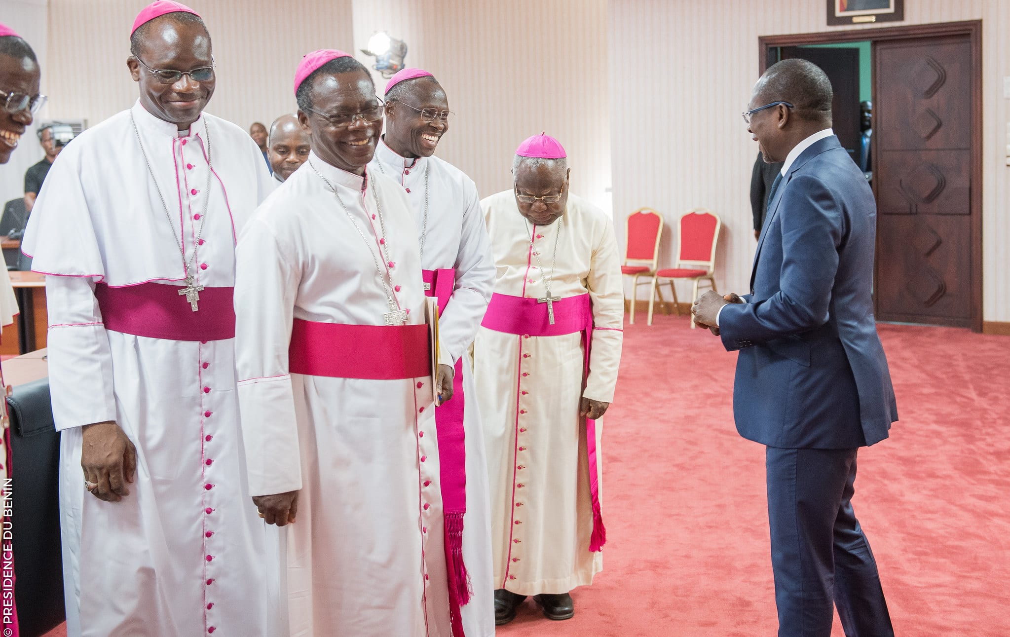 Quatre ecclésiastiques en habits liturgiques et un homme en costume discutent dans une salle avec tapis rouge et chaises rouges vides.