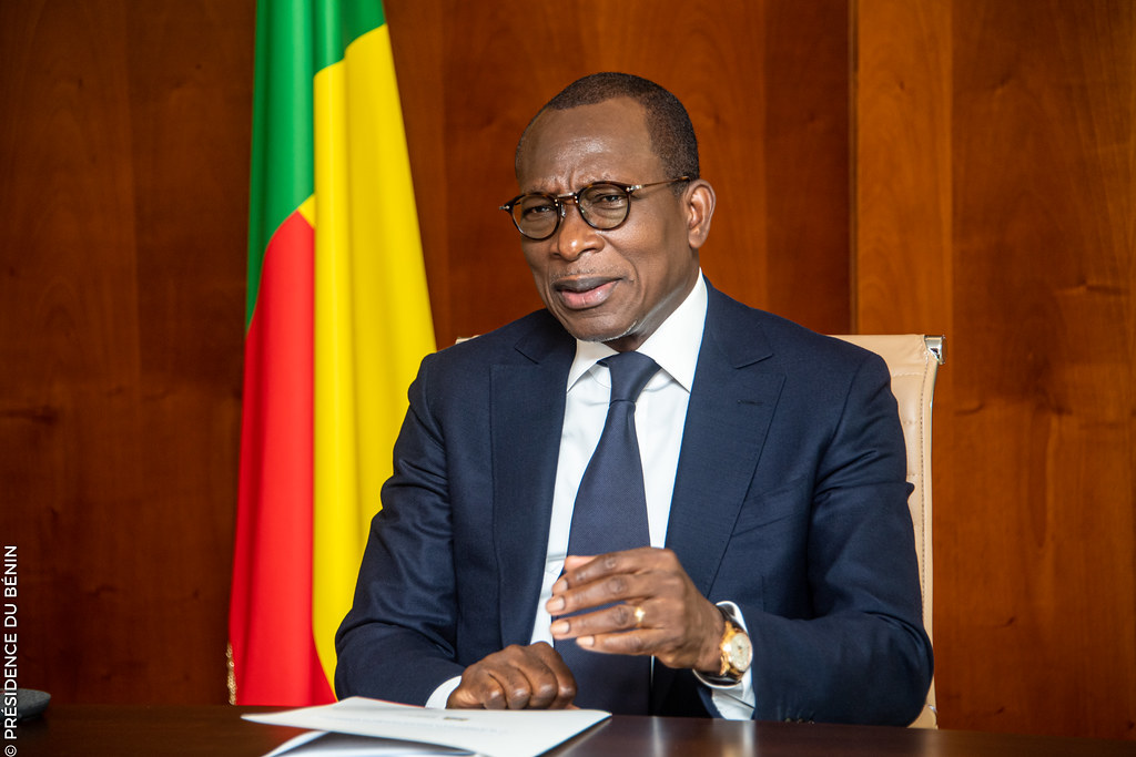 Homme en costume sombre et lunettes parlant assis devant un drapeau tricolore vert, jaune et rouge sur fond bois