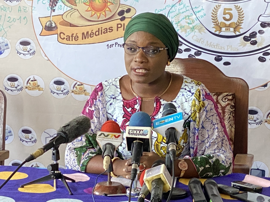 Femme en tenue colorée et foulard vert qui parle face à plusieurs micros lors d’une conférence de presse