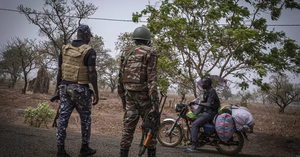 Deux soldats armés parlent à un homme assis sur une moto chargée de sacs sur une route bordée d’arbres en zone rurale