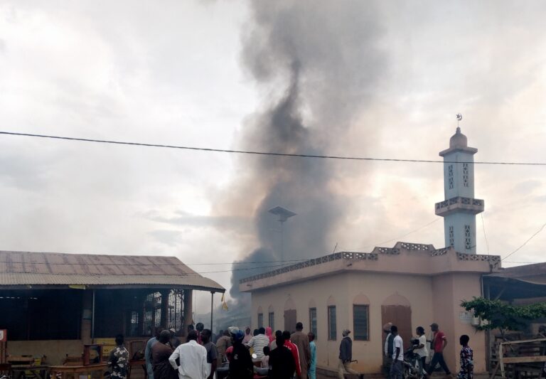 Des habitants regardent une épaisse fumée noire s'élever derrière un bâtiment à côté d'une mosquée avec un ciel nuageux