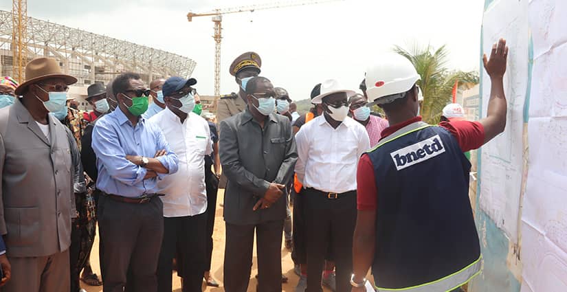 Un groupe d’hommes en tenue formelle et casques observe un plan affiché sur un chantier avec une grue en arrière-plan