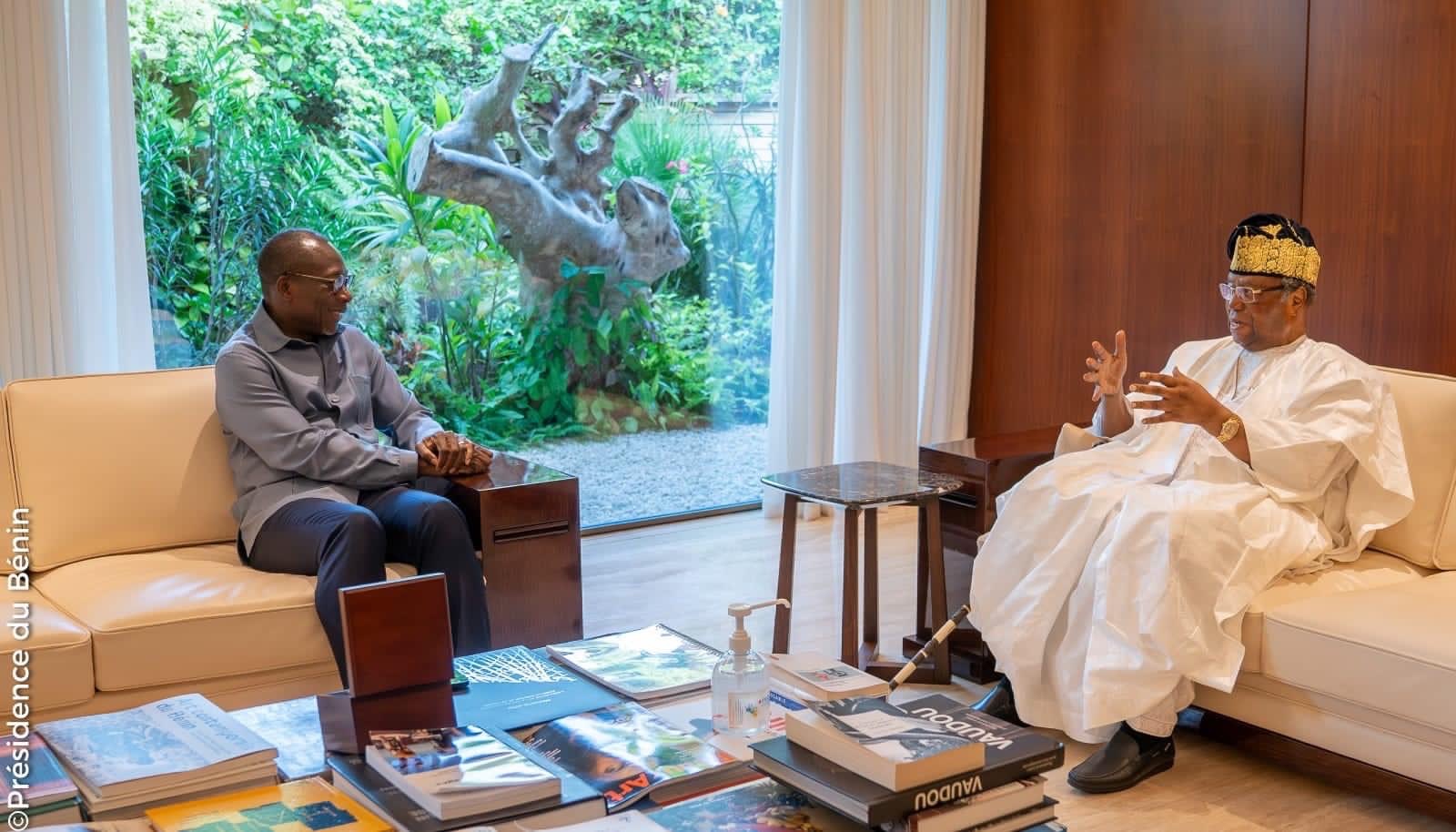 Deux hommes discutent assis dans un salon moderne avec une grande fenêtre donnant sur un jardin et des livres sur une table basse
