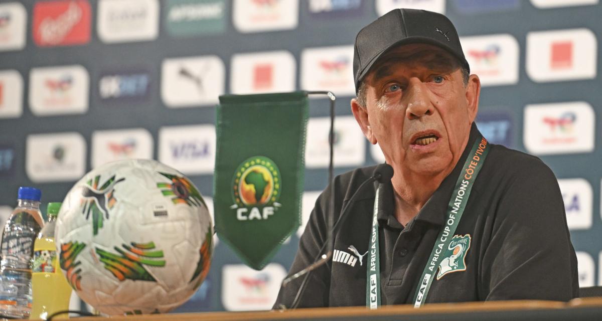 Homme portant une casquette noire parle à une conférence de presse avec un ballon et un fanion de football devant lui