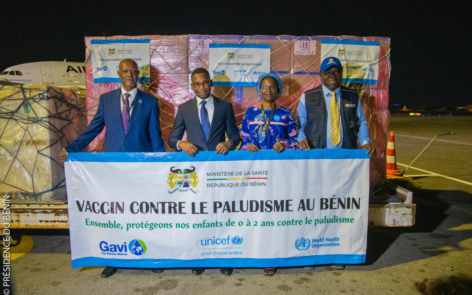 Quatre personnes tiennent une banderole devant des palettes de cartons empilés et un avion de nuit