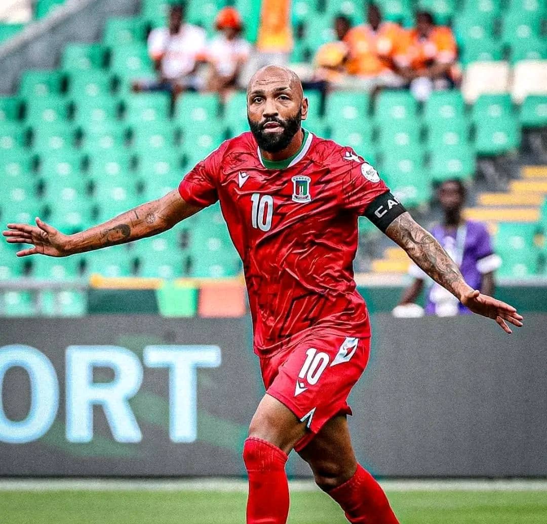 Un footballeur en maillot rouge numéro 10 célèbre un but sur un terrain avec gradins verts en arrière-plan