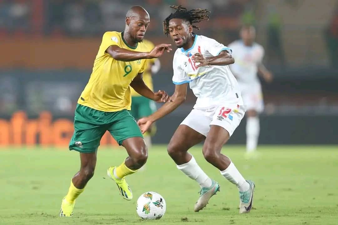 Deux joueurs de football s'affrontent sur le terrain, l'un en jaune et vert, l'autre en blanc et bleu, près du ballon sur le gazon