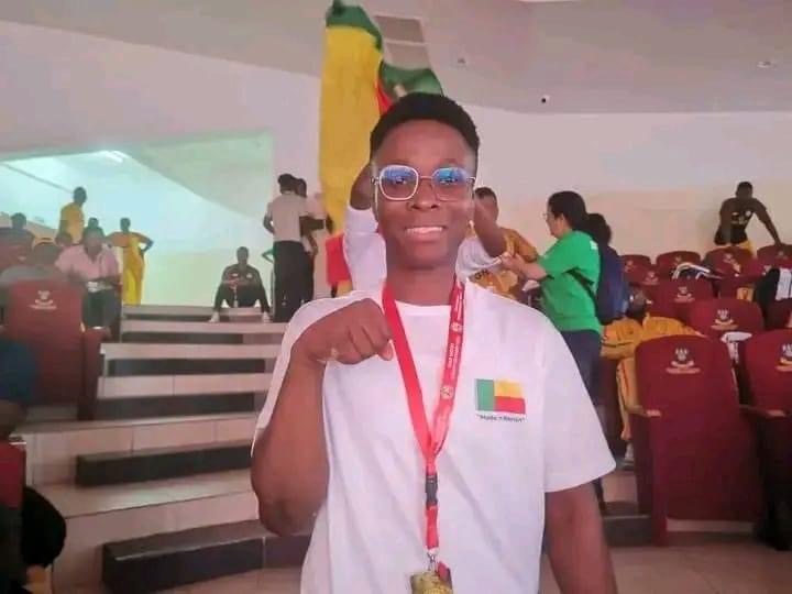 Jeune homme souriant avec médaille autour du cou et drapeau coloré en arrière-plan dans une salle avec gradins