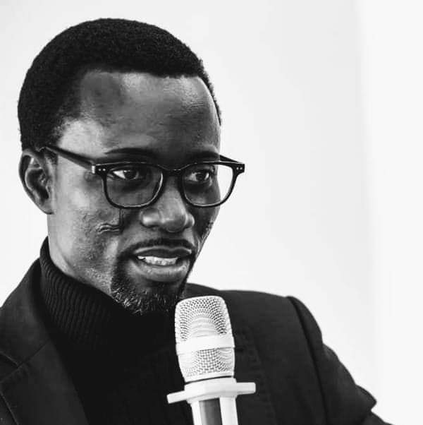 Homme portant des lunettes et un pull noir parlant dans un micro blanc sur fond clair