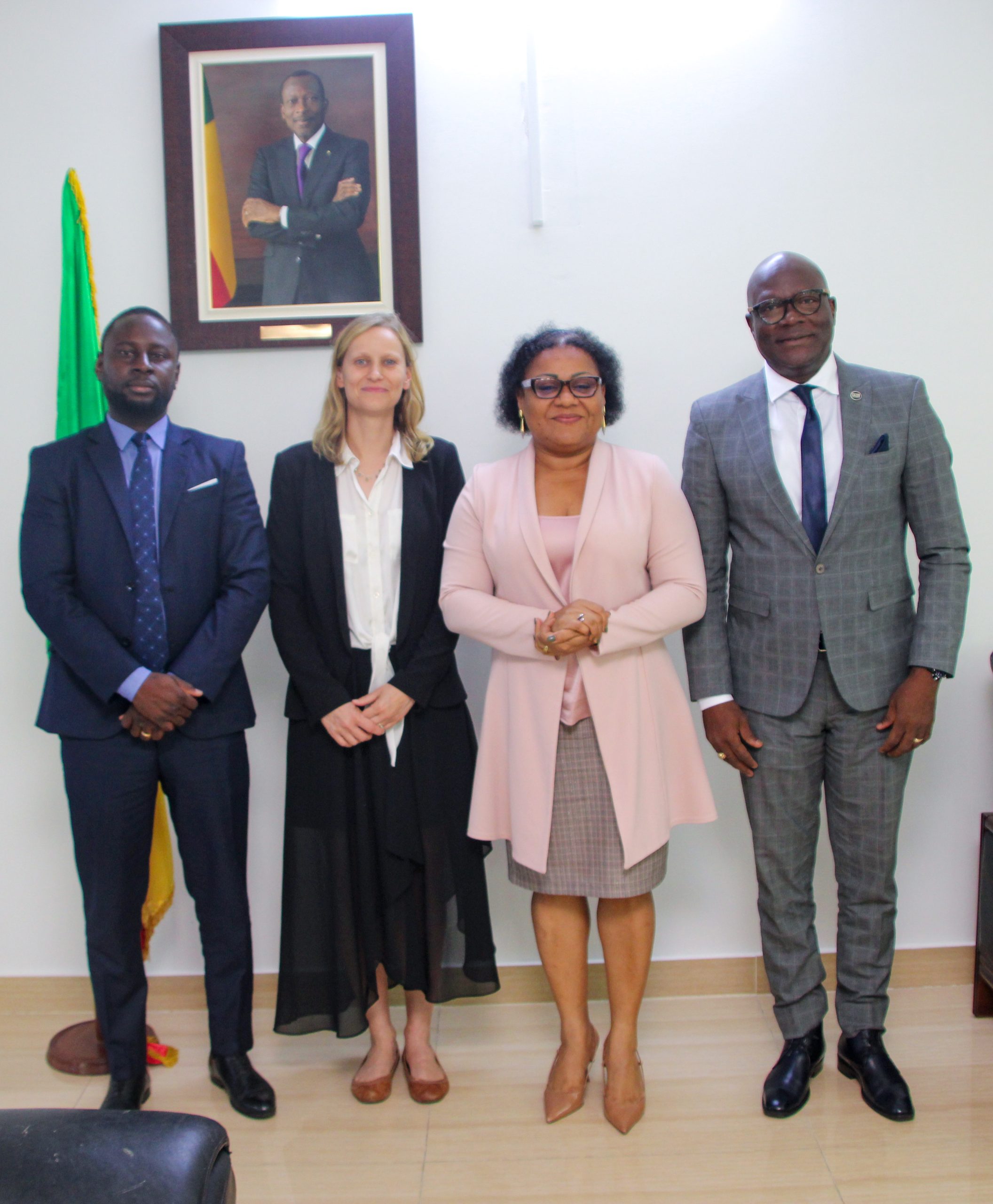 Quatre personnes en tenue professionnelle posent debout devant un mur blanc avec un portrait encadré et un drapeau partiellement visible à gauche