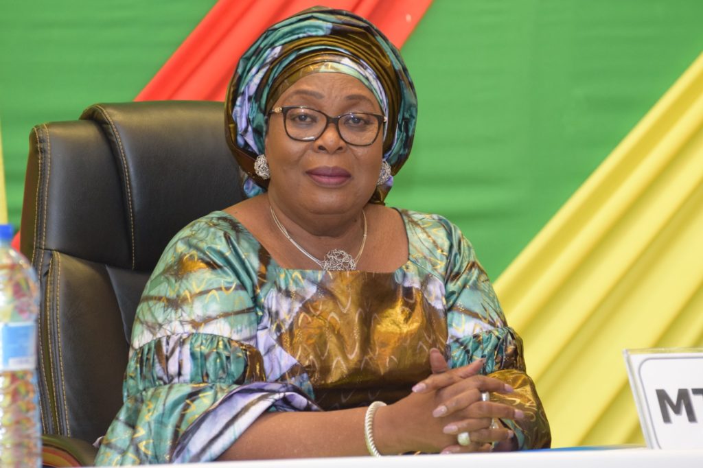 Femme assise en robe colorée et turban assorti devant un fond vert, rouge et jaune avec une bouteille d'eau et un panneau partiel MTN visible