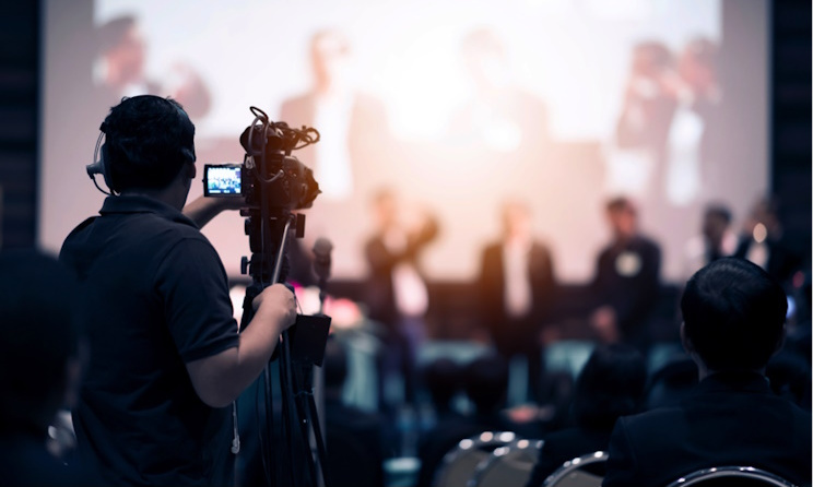 Une personne filme une conférence où plusieurs intervenants sont assis devant un public dans une salle obscure
