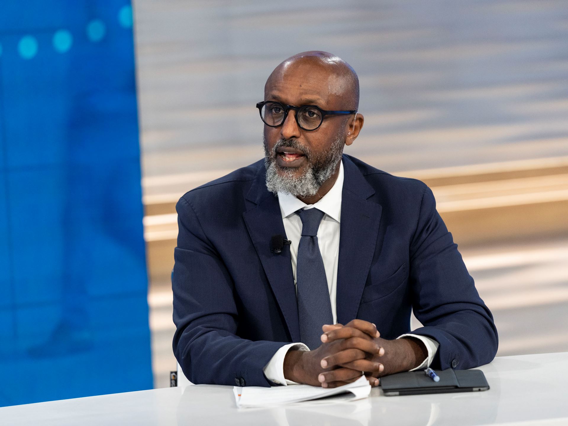 Homme d’affaires portant un costume sombre et des lunettes, assis à une table avec un carnet et un stylo devant lui