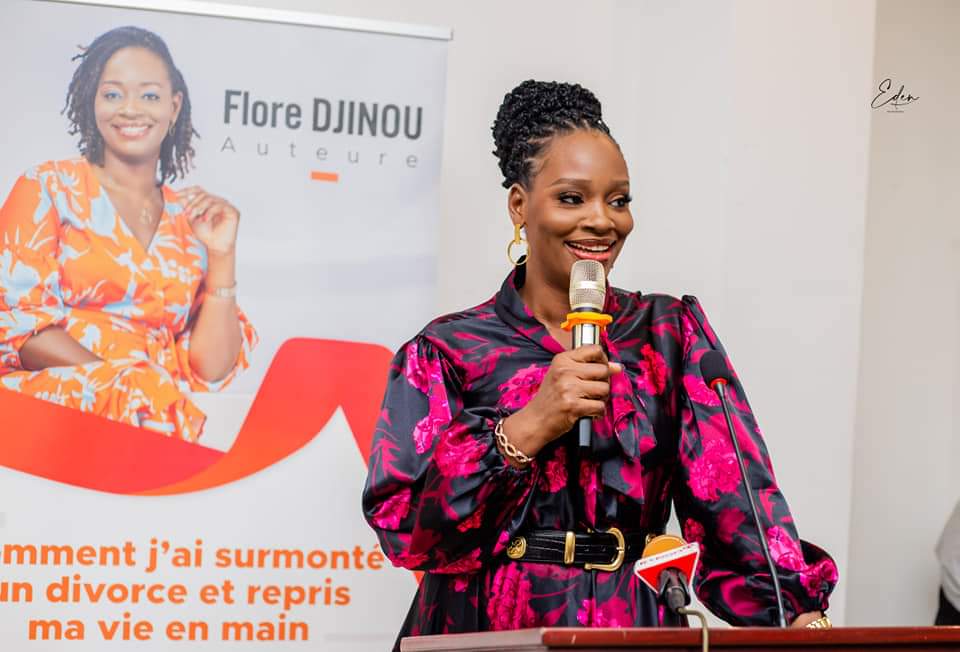 Femme souriante parlant au micro lors d'une conférence avec un poster promotionnel en arrière-plan.