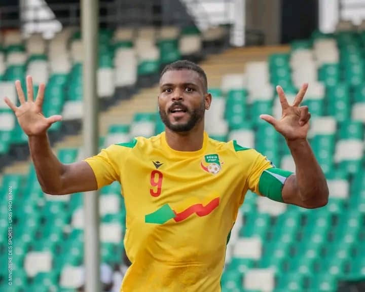 Joueur de football en maillot jaune numéro 9 et brassard vert levant les deux mains dans un stade aux sièges verts et blancs