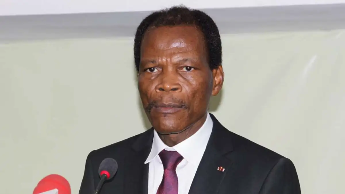 Homme en costume sombre et cravate donnant un discours devant un micro dans une salle neutre