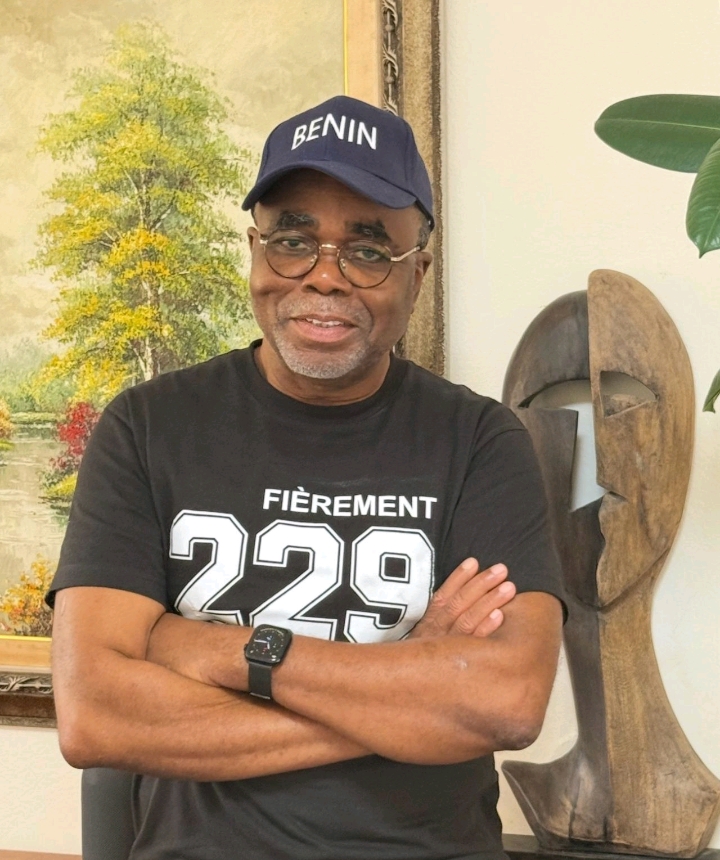 Homme souriant portant une casquette bleue avec mention BENIN et un t-shirt noir marqué FIÈREMENT 229 bras croisés devant un tableau et une sculpture en bois