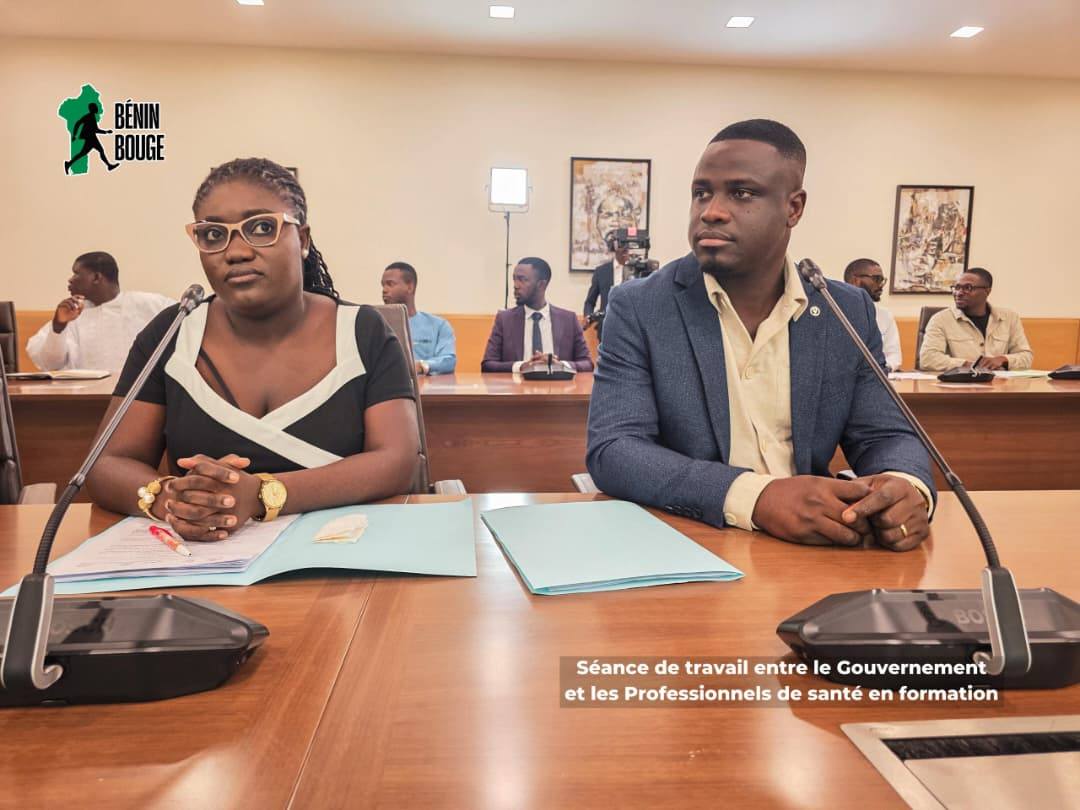 Deux personnes assises à une table lors d'une séance de travail dans une salle de réunion avec plusieurs autres participants en arrière-plan