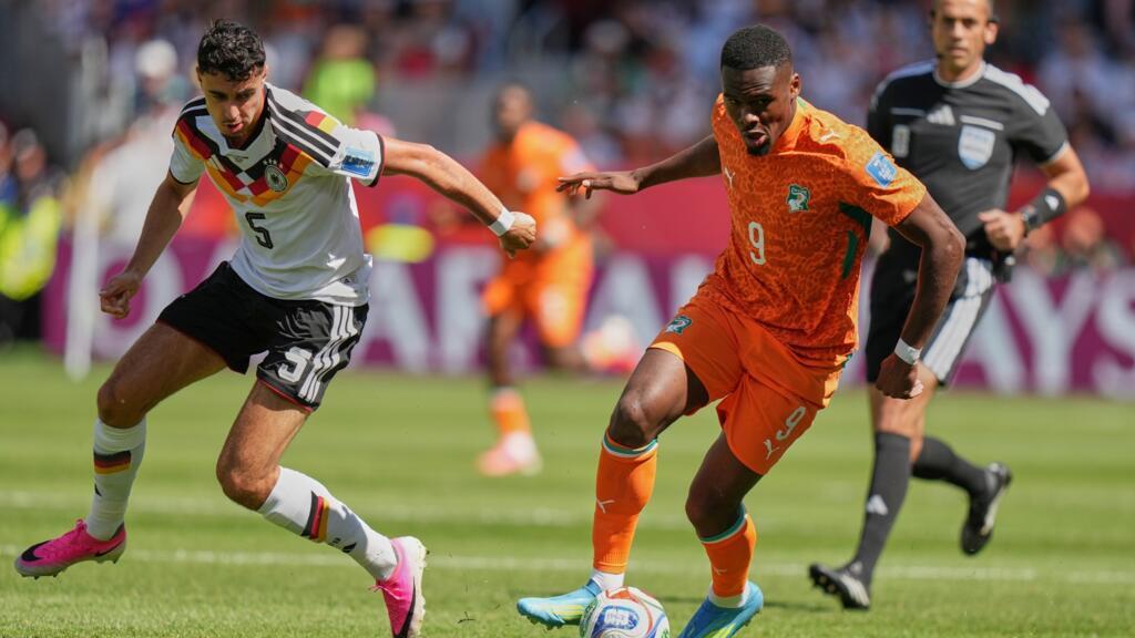 Deux joueurs en action lors d’un match de football, l’un en maillot blanc et l’autre en maillot orange, avec un arbitre en arrière-plan