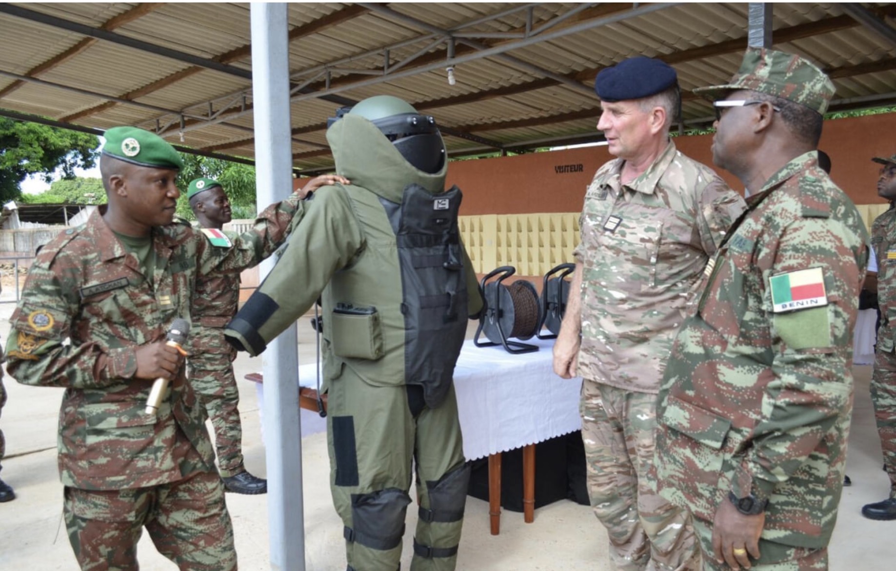 Des militaires en uniforme examinent une tenue de déminage verte sous un abri avec un mobilier blanc en arrière-plan